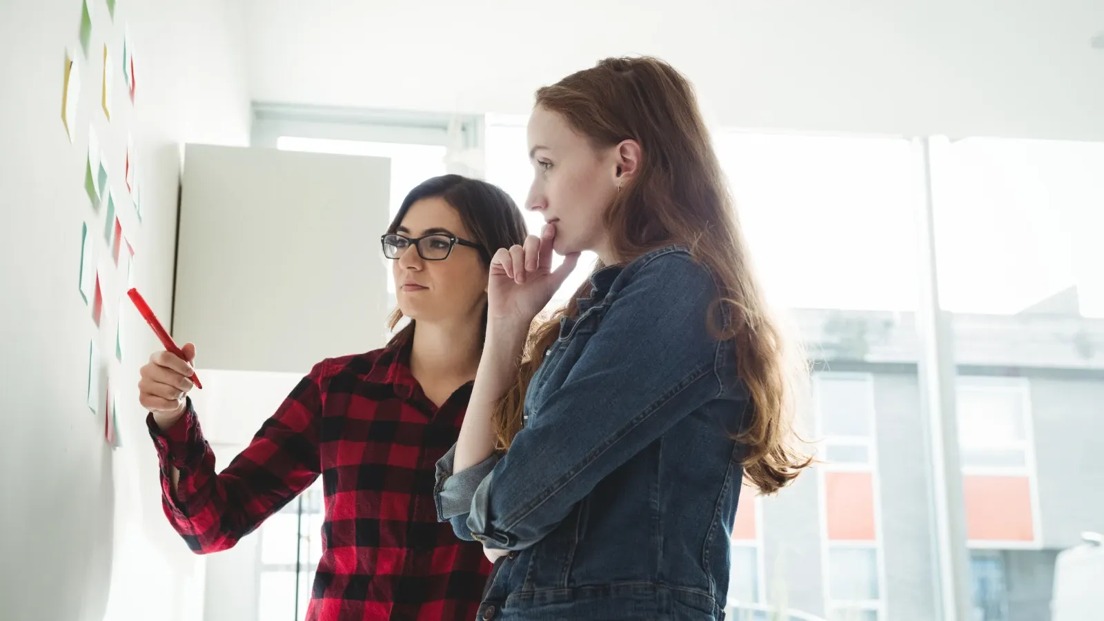 Zwei Frauen stehen neben einer weißen Tafel, eine zeigt mit einem roten Stift auf Haftnotizen, während die andere nachdenklich in einem hell erleuchteten Raum beobachtet.